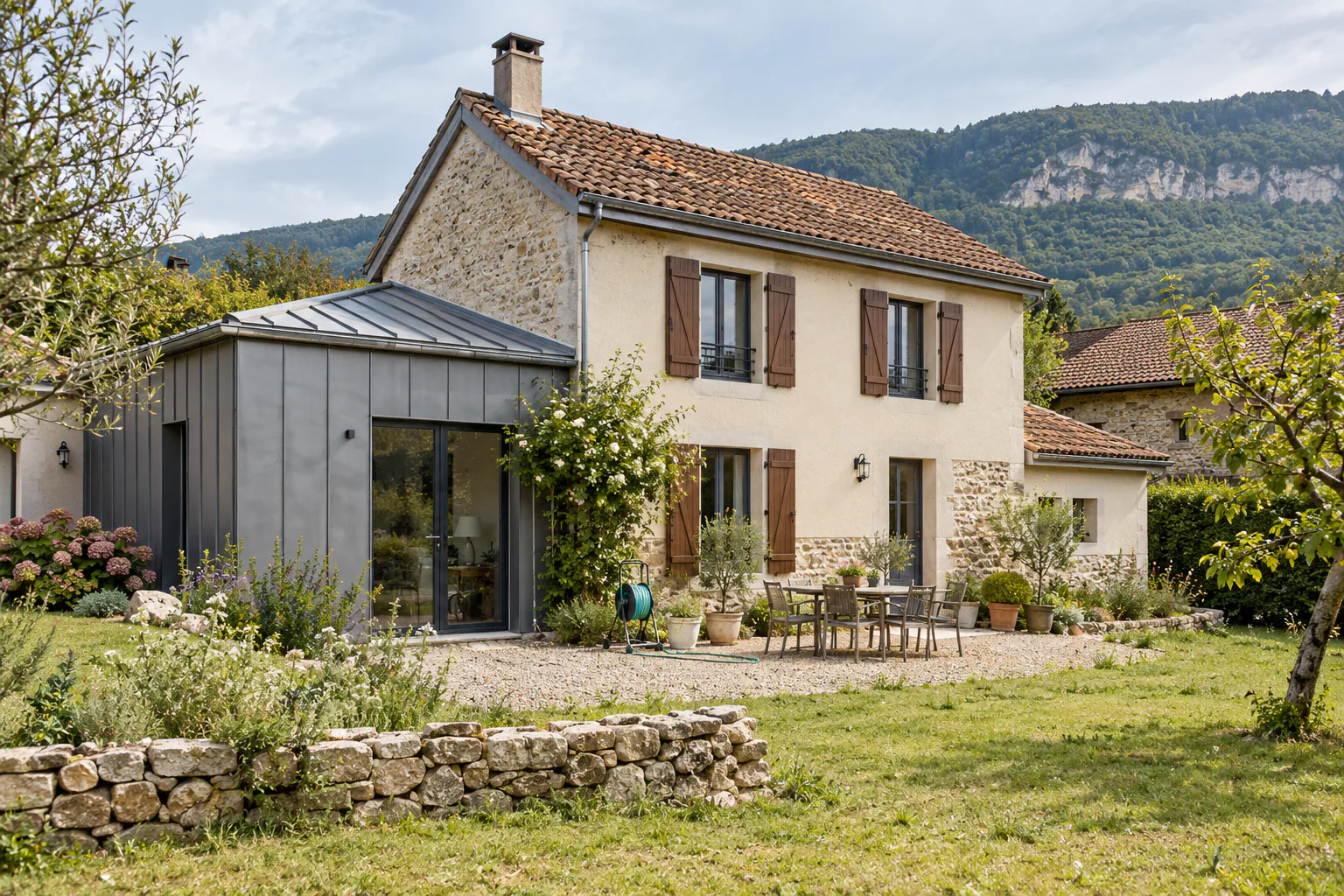 Maison ancienne avec petite extension métallique Une maison aux volets bruns s’ouvre sur un jardin, avec une petite extension latérale à toit métallique.