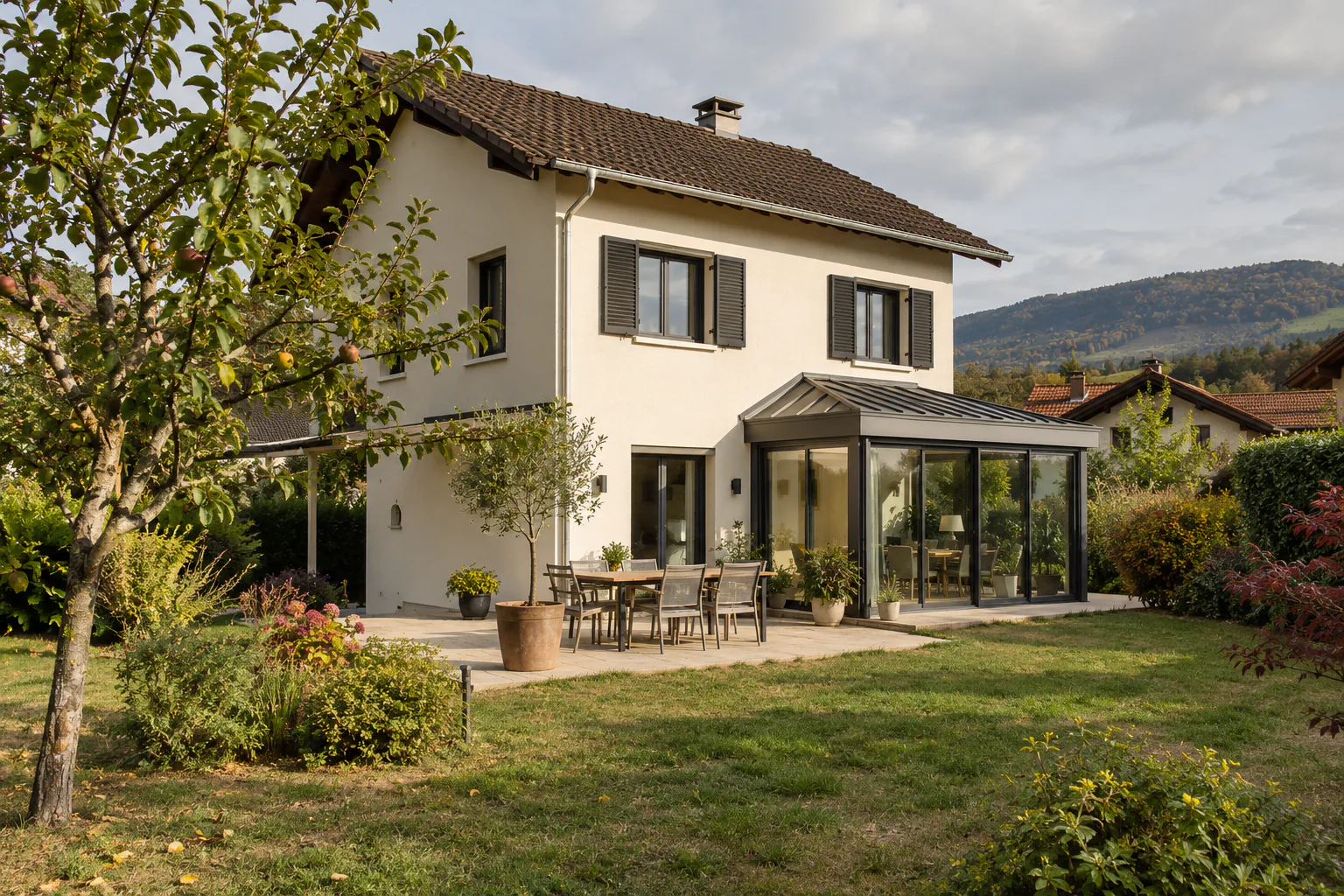 Maison claire et extension vitrée sur terrasse Une petite extension vitrée à toiture inclinée s’adosse à une maison claire, devant une terrasse avec table.