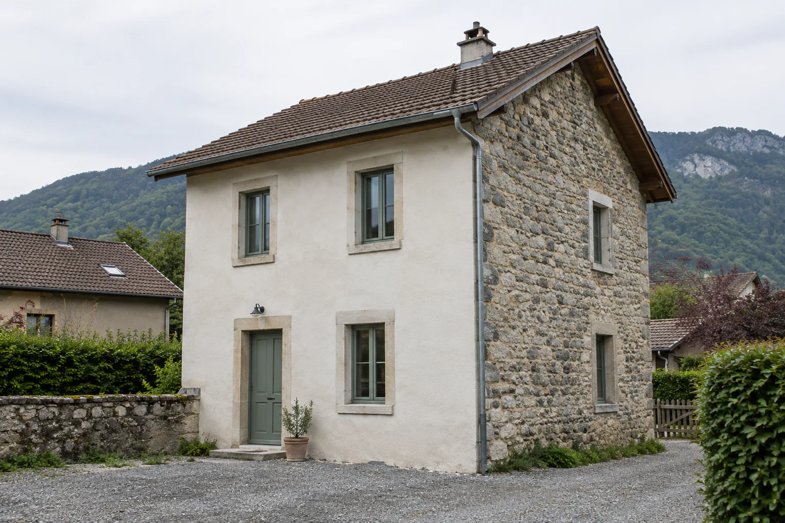 Maison étroite avec pignon en pierre apparente Une petite maison à étage associe une façade claire et un pignon en pierre, devant une cour de gravier.