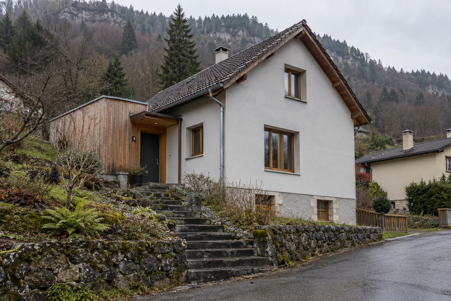 Maison blanche et extension bois sur terrain pentu Un escalier extérieur en pierre mène à une maison blanche et à son extension en bois, au-dessus de la route.
