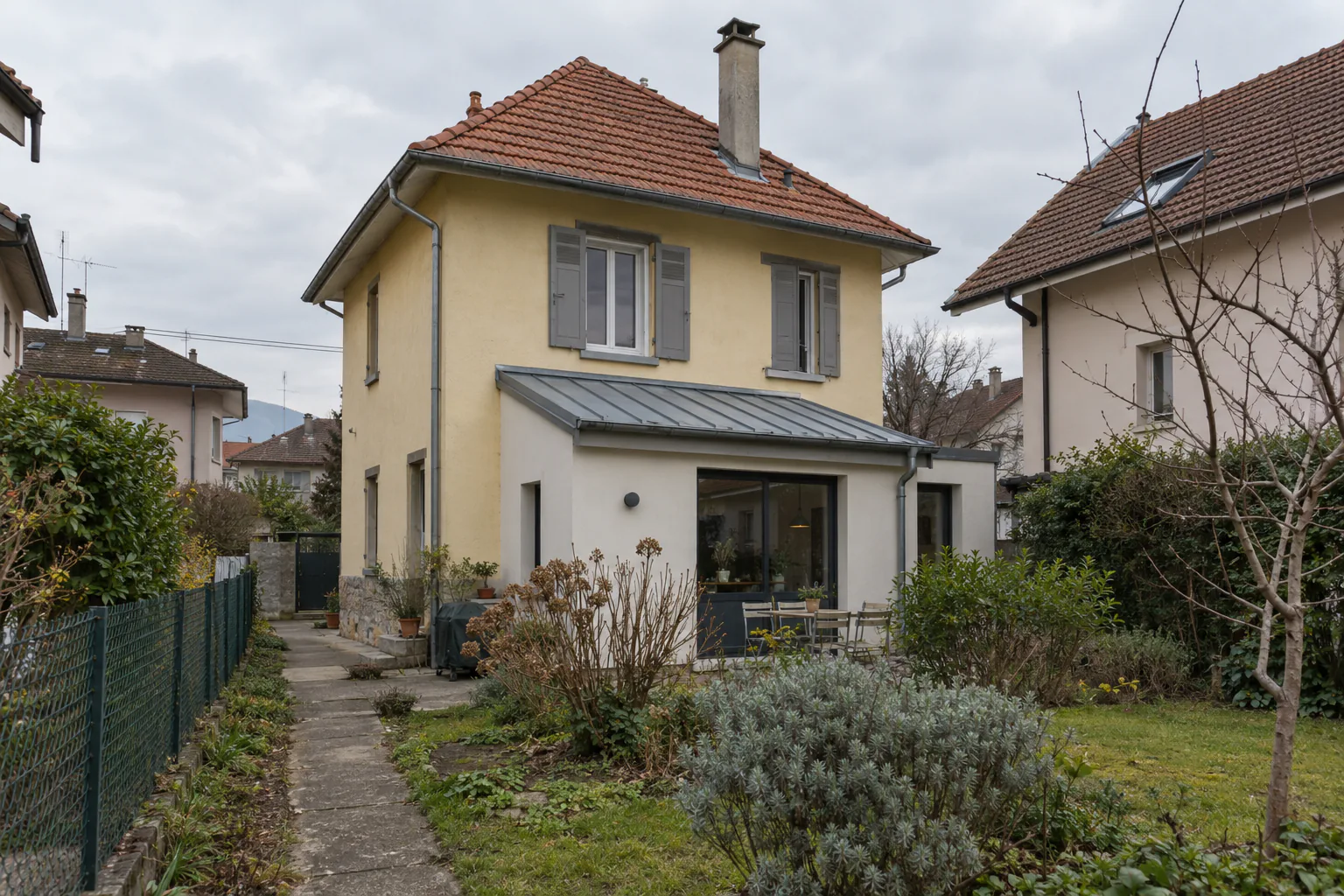 Petite maison jaune et extension sur jardin Une maison jaune à étage possède une extension claire à baie vitrée, dans un jardin étroit entre voisins.