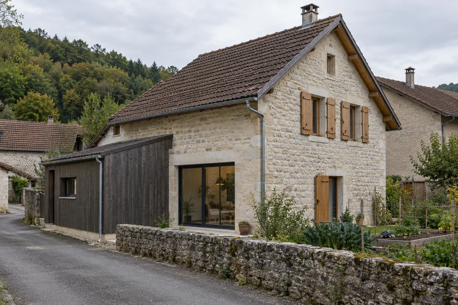 Maison de pierre et extension en bois sombre Une maison aux volets bruns est complétée par un petit volume en bois sombre, le long d’une rue et d’un muret.