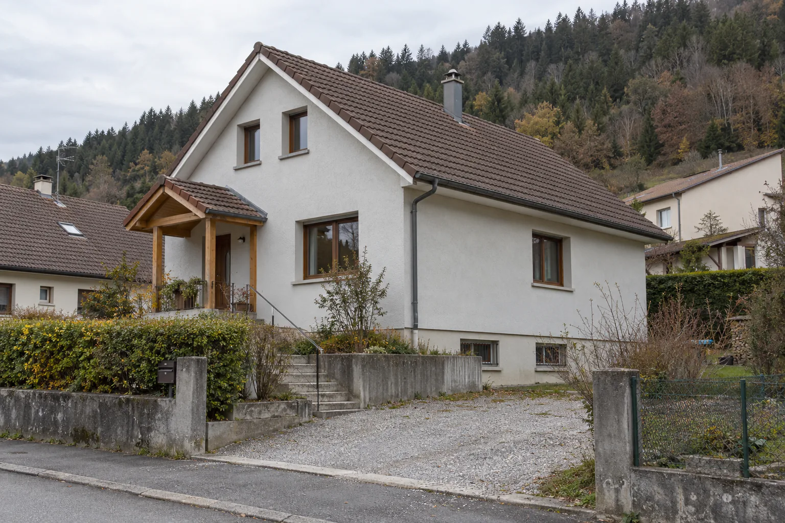 Maison claire avec petit porche en bois Une maison blanche à toit pentu présente un petit porche en bois, derrière une allée et un muret de pierre.