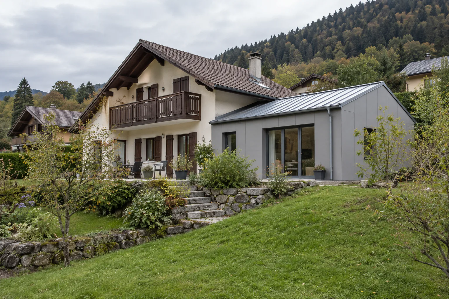 Maison à balcon et extension blanche en pente Une extension blanche à toiture métallique prolonge une maison avec balcon, devant des marches en pierre et une pelouse.