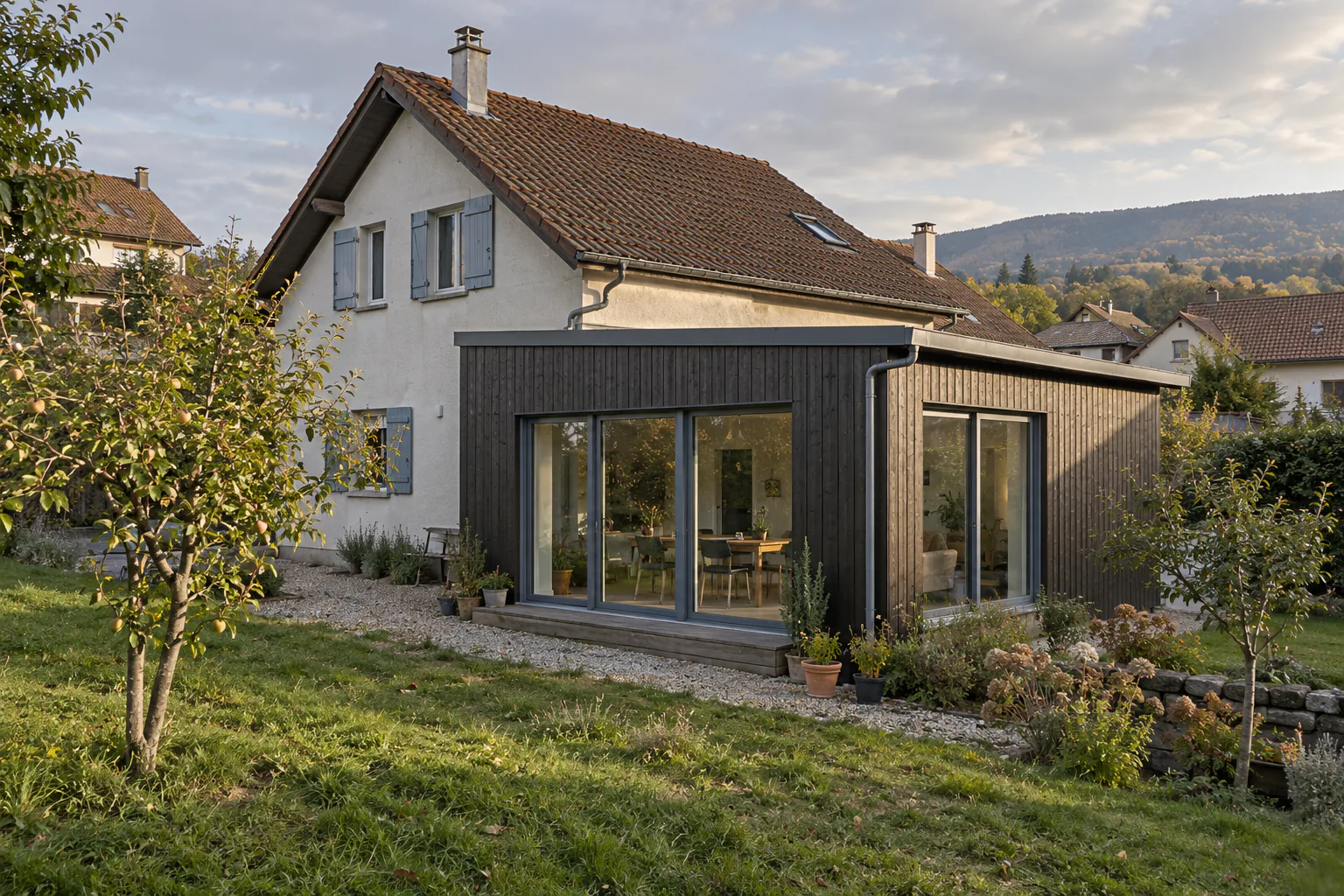 Extension en bois sombre ouverte sur terrasse Une extension basse habillée de bois sombre présente deux façades vitrées, devant une terrasse et un jardin.