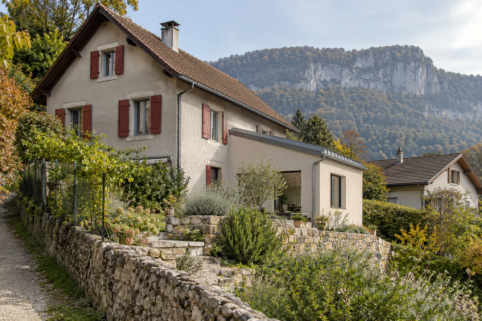 Maison aux volets rouges sur jardin en pente Une maison ancienne aux volets rouges et à petite extension claire domine un jardin bordé de murs en pierre.