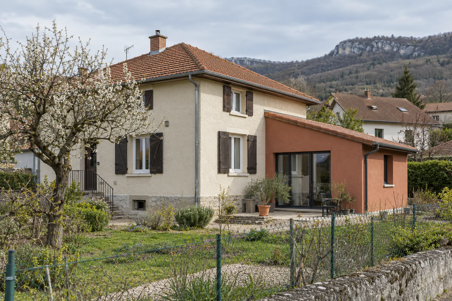 Maison claire et extension en enduit orangé Une extension orangée à baie vitrée s’adosse à une maison claire, derrière un potager et une clôture.