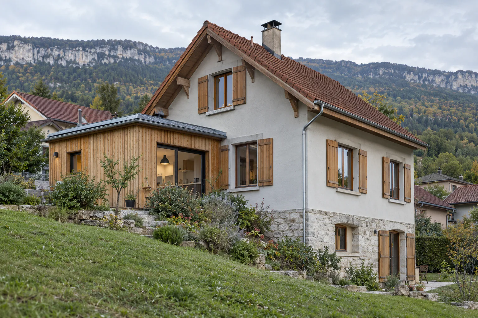 Maison sur sous-sol et extension bois en pente Une petite extension bois vitrée rejoint une maison claire au soubassement de pierre, devant une pelouse en pente.