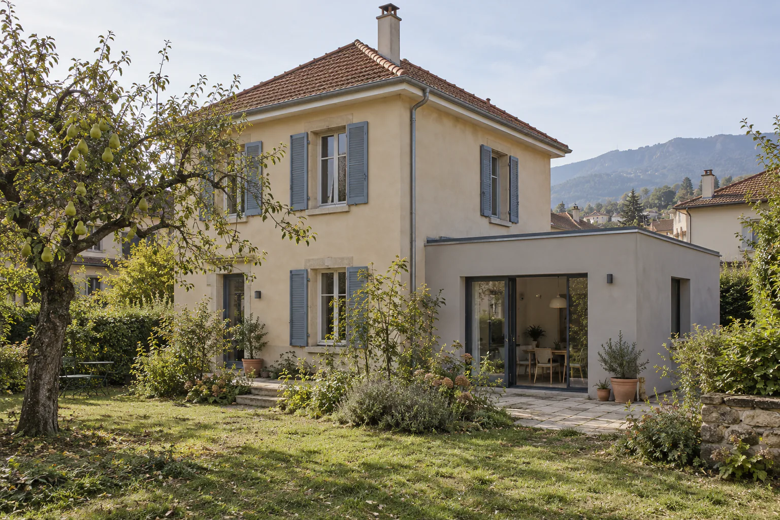 Maison claire et extension grise sous un arbre Une extension grise à grande porte vitrée s’adosse à une maison à étage, devant un arbre et une pelouse.