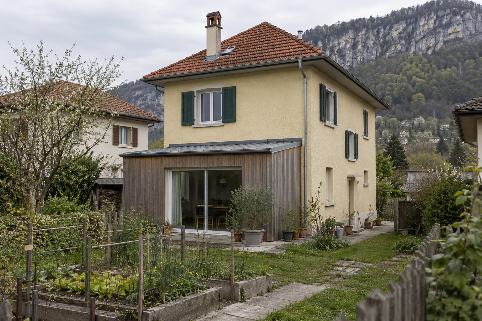 Maison haute et extension bois devant le potager Une extension basse en bois complète une maison à trois niveaux, devant une pelouse et un petit potager clôturé.