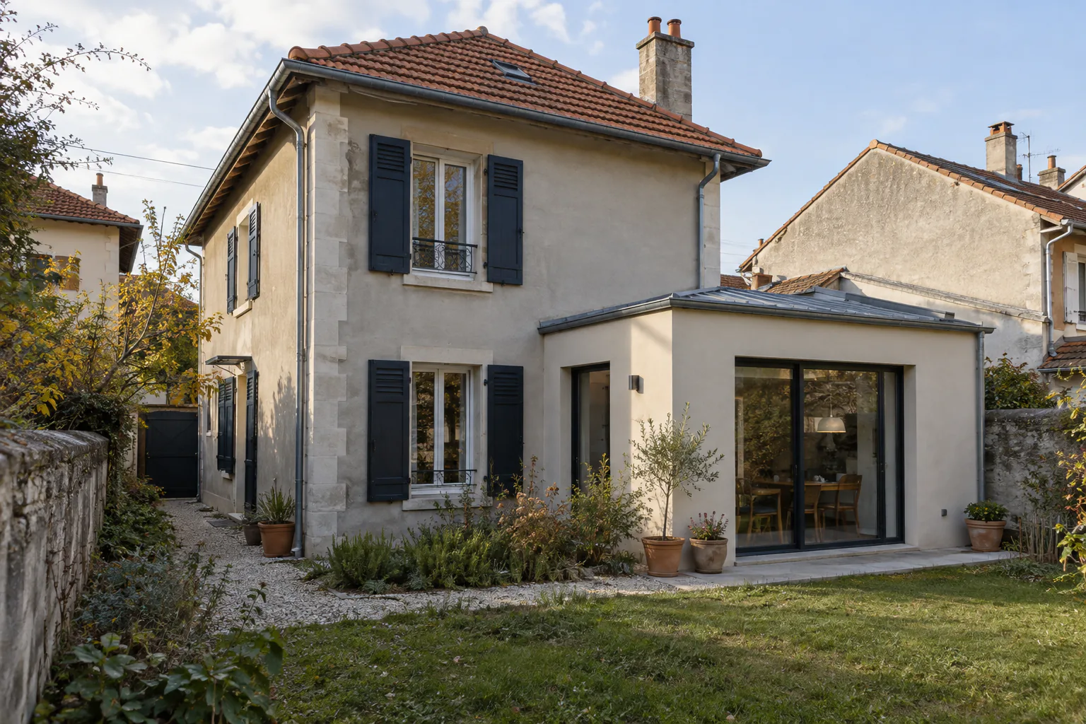 Maison aux volets sombres et extension vitrée Une extension claire à grande baie vitrée prolonge une maison aux volets bleu sombre, dans un jardin bordé de murs.