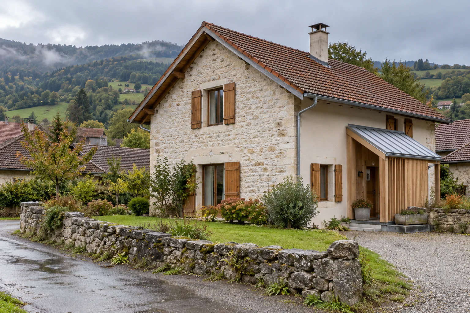 Maison de pierre et extension bois près de la route Une maison à pignon en pierre possède une petite extension bois à toit métallique, derrière un muret bordant la route.