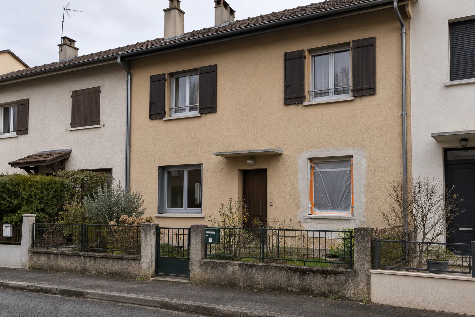 Maison mitoyenne aux volets bruns sur rue Une maison mitoyenne à enduit ocre et volets bruns donne sur la rue, derrière une petite clôture métallique.