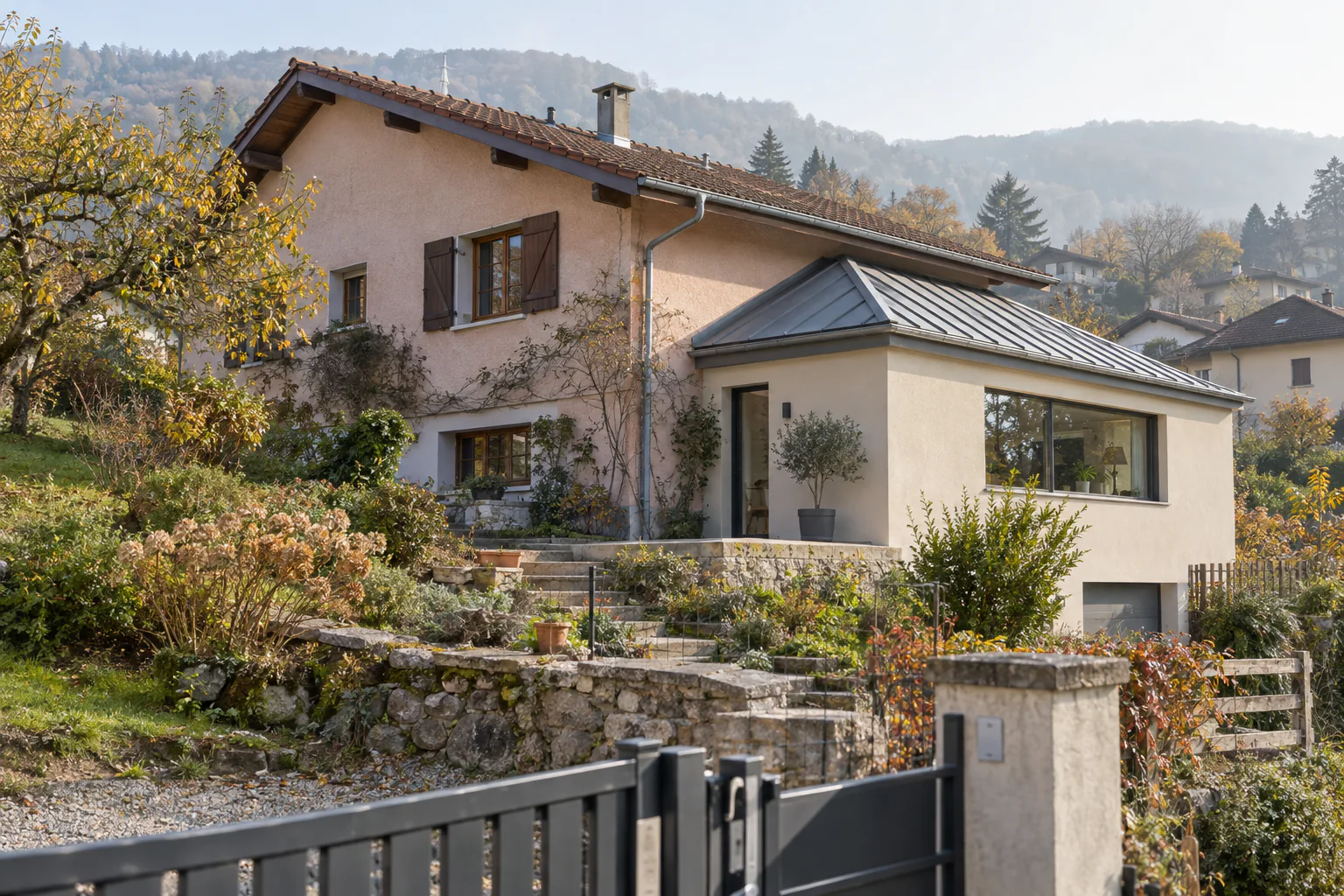 Maison rose et extension claire à toiture métallique Une extension claire à trois fenêtres et toiture métallique prolonge une maison rose, derrière un muret en pierre.