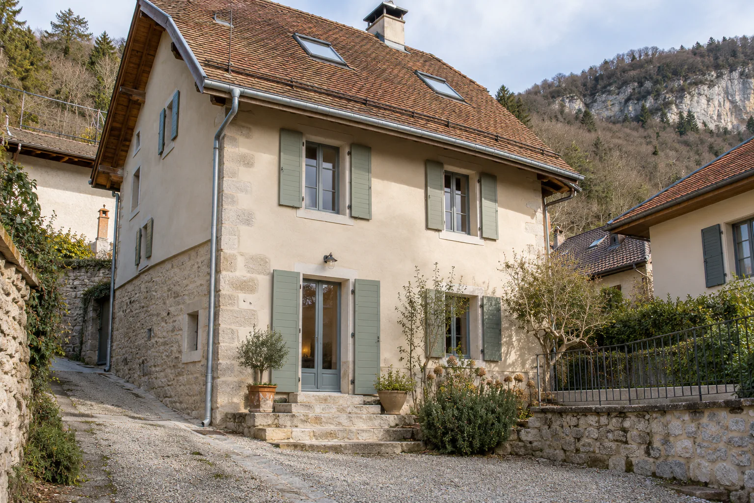 Maison ancienne et fenêtres de toit dans une cour Une maison à enduit patiné, volets gris et soubassement de pierre possède deux fenêtres de toit au-dessus d’une cour.