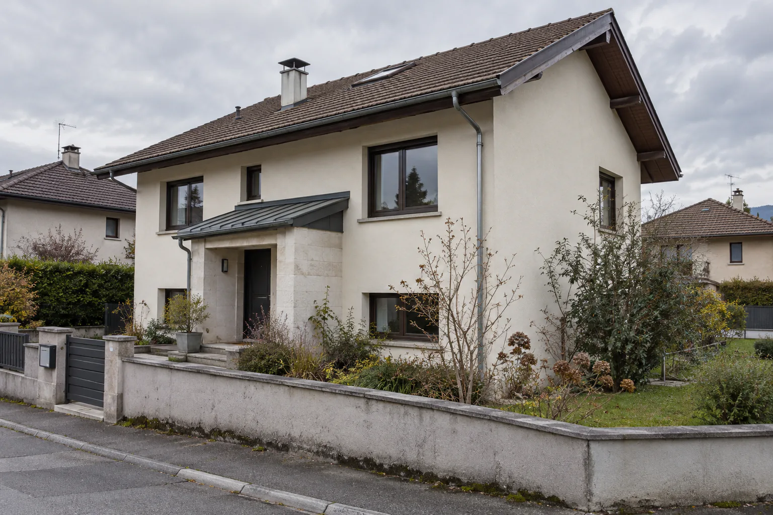Entrée couverte sur une maison claire à étage Une petite toiture métallique protège l’entrée d’une maison claire, derrière un muret enduit et un jardin.