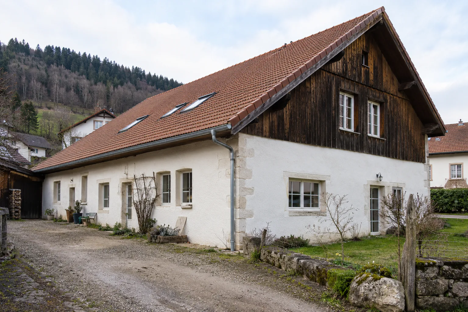 Longue maison et grand pignon en bois sombre Une longue maison blanche présente un pignon supérieur en bois sombre et des fenêtres de toit, le long d’une allée.