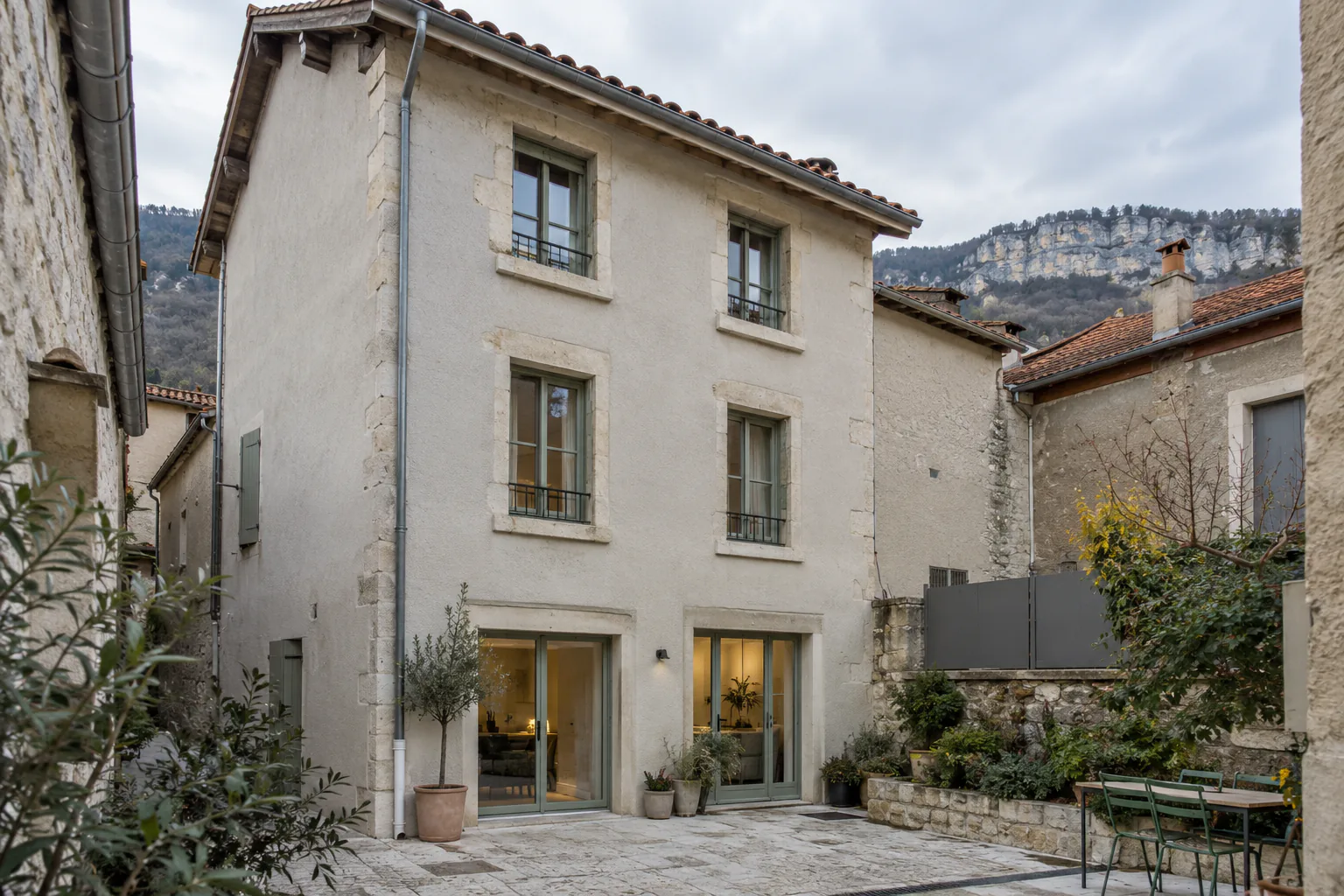 Maison étroite et terrasse dans une cour close Une maison étroite à trois niveaux s’ouvre au rez-de-chaussée sur une cour pavée avec table et chaises.