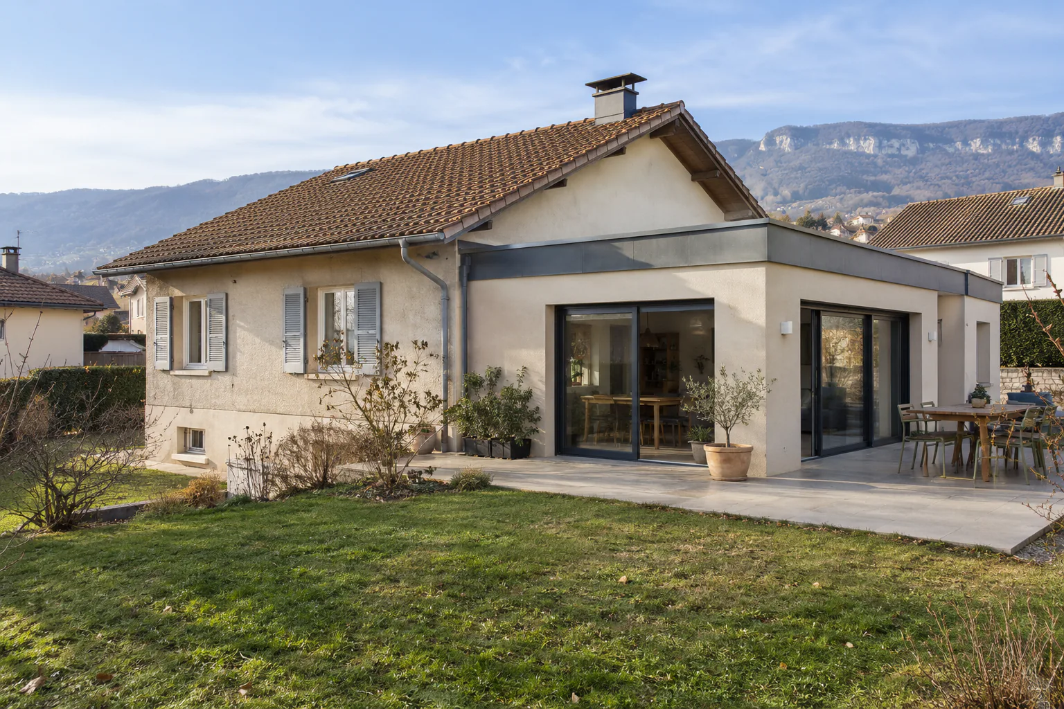 Pavillon et extension minérale largement vitrée Une extension à revêtement minéral et larges baies vitrées prolonge un pavillon, au bord d’une terrasse et d’une pelouse.