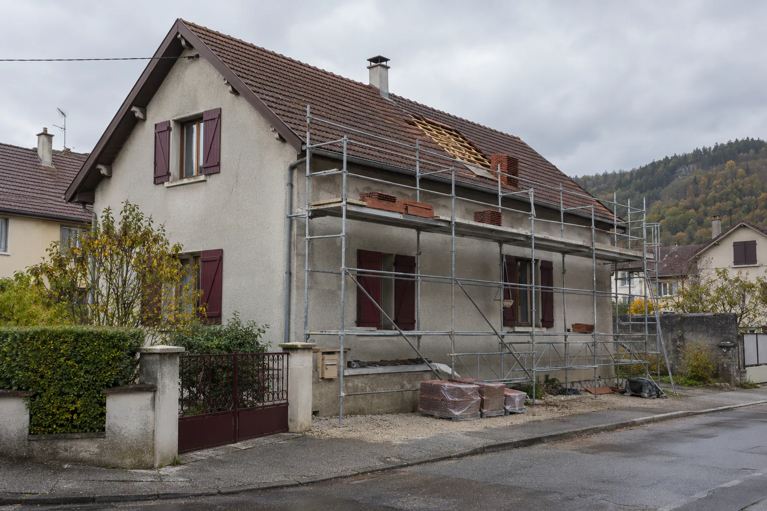 Échafaudage devant une façade en rénovation Un échafaudage couvre la façade d’une maison aux volets bruns, avec matériaux et palettes rangés au pied du mur.