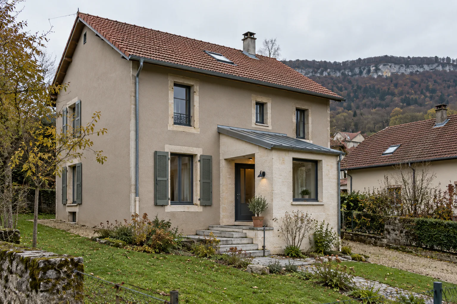Maison ancienne et petit volume d’entrée neuf Une petite extension claire à toit métallique forme l’entrée d’une maison aux volets gris, au-dessus d’un jardin.