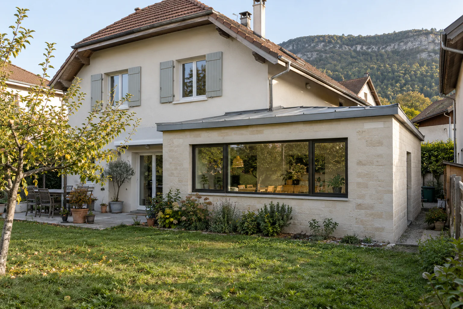 Maison à volets gris et large extension blanche Une extension blanche à toit bas présente une grande fenêtre horizontale devant le jardin d’une maison à étage.