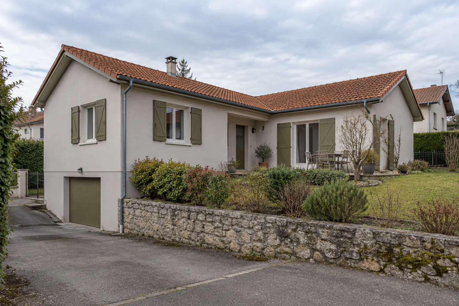Maison basse sur garage et muret en pierre Une maison claire en deux volumes sous tuiles possède un garage en contrebas et un jardin retenu par un mur en pierre.