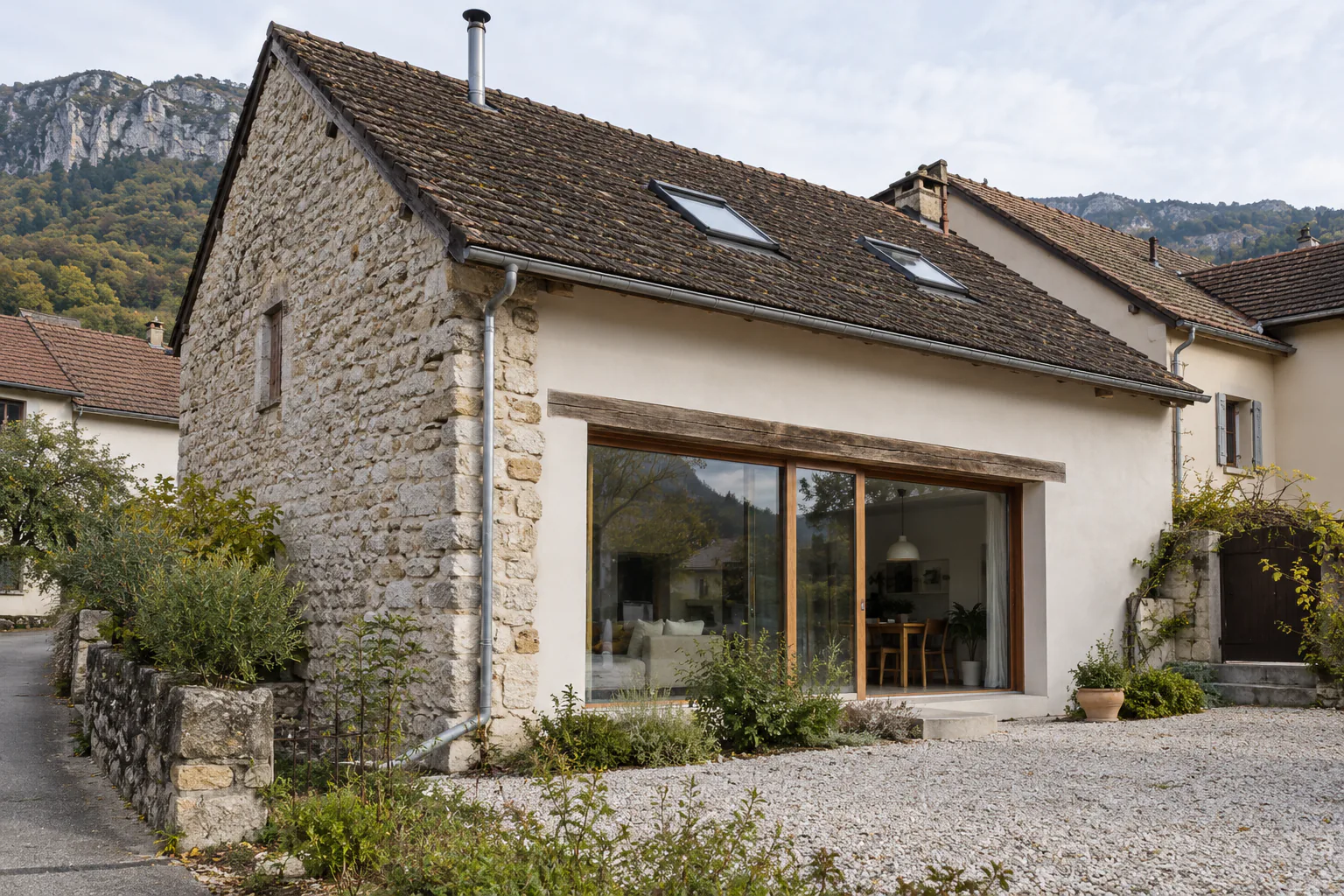 Petite grange de pierre ouverte sur une cour Une grande baie vitrée sous une poutre en bois ouvre le pignon d’une petite grange en pierre sur une cour de gravier.