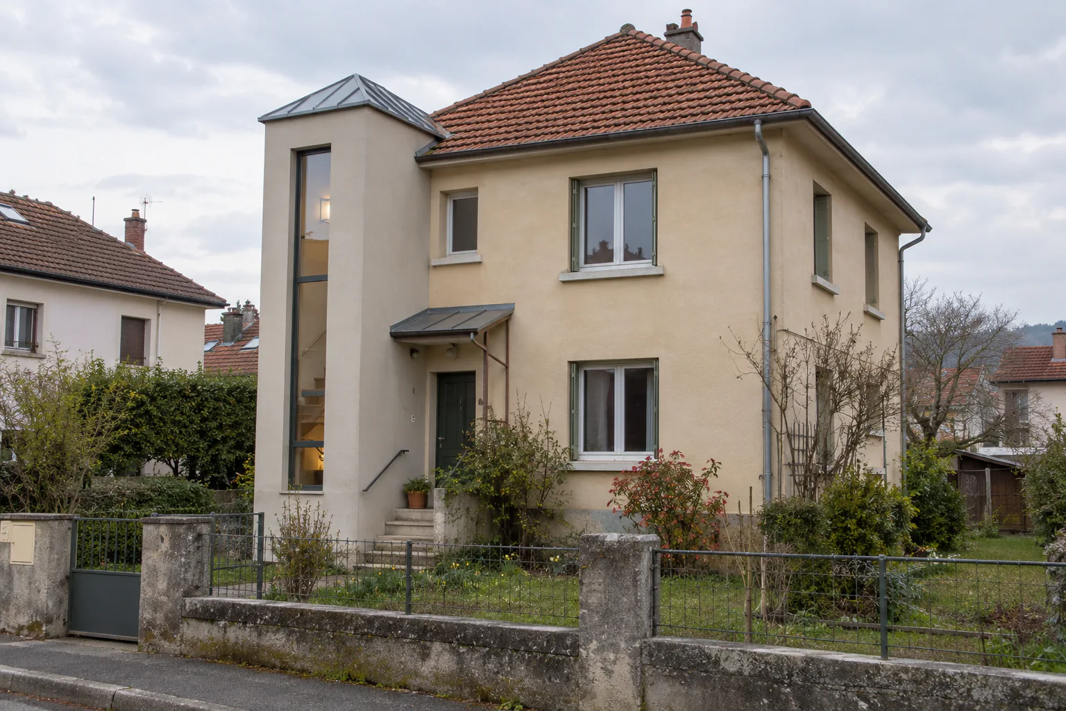 Maison à étage avec haute cage d’entrée claire Un volume vertical clair à fenêtre étroite se détache contre une maison à toiture rouge, derrière une clôture basse.