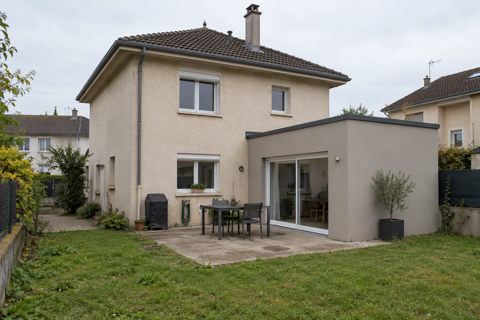 Maison à toit brun et extension grise sur terrasse Une extension grise à toit plat et porte vitrée s’adosse à une maison claire, devant une terrasse avec table.