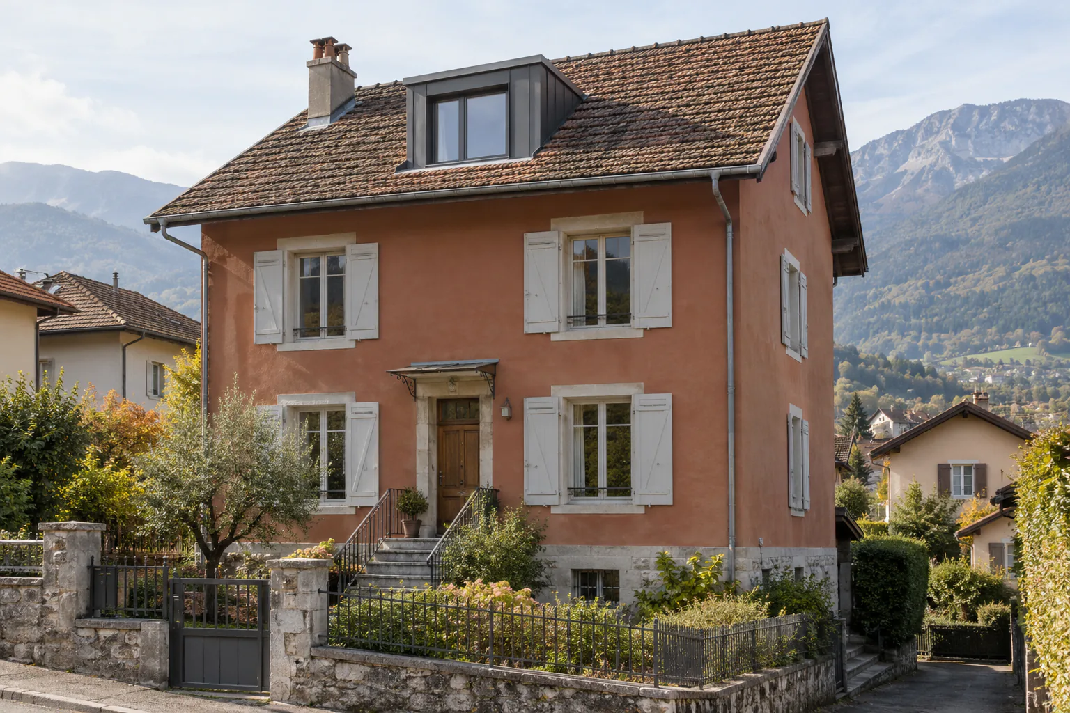 Maison rose avec lucarne et volets clairs Une maison rose à étage possède une lucarne et des volets clairs, derrière un jardin et une clôture sur muret.