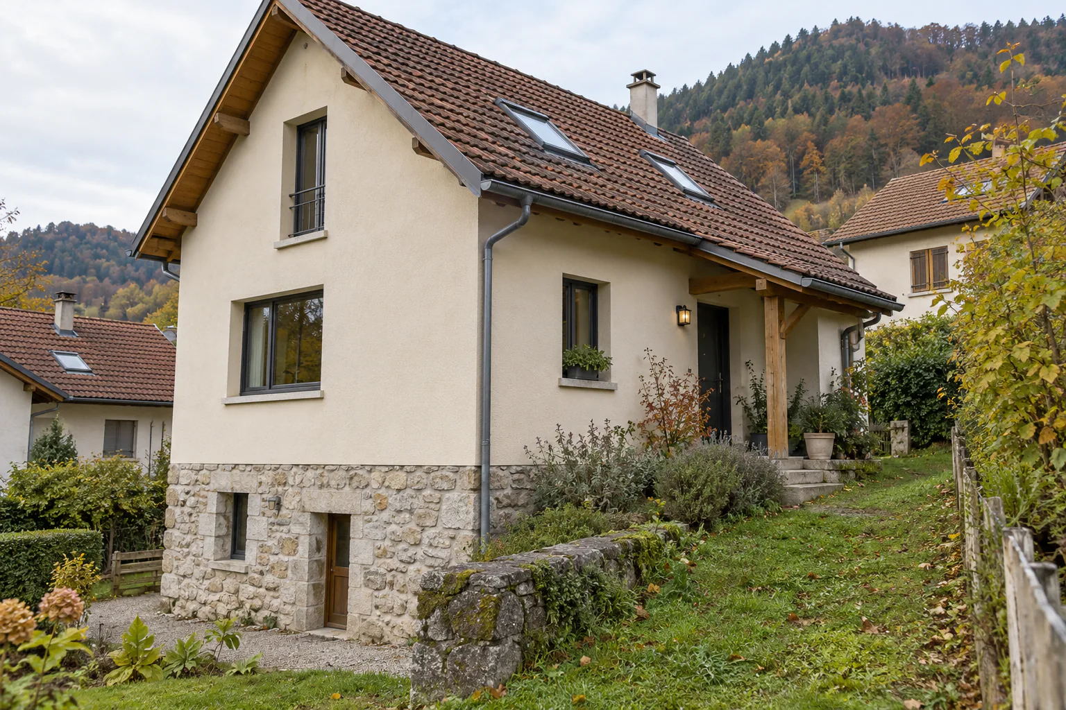 Maison sur soubassement de pierre en pente Une maison claire à toiture percée de fenêtres de toit s’élève sur un soubassement de pierre, dans un jardin pentu.