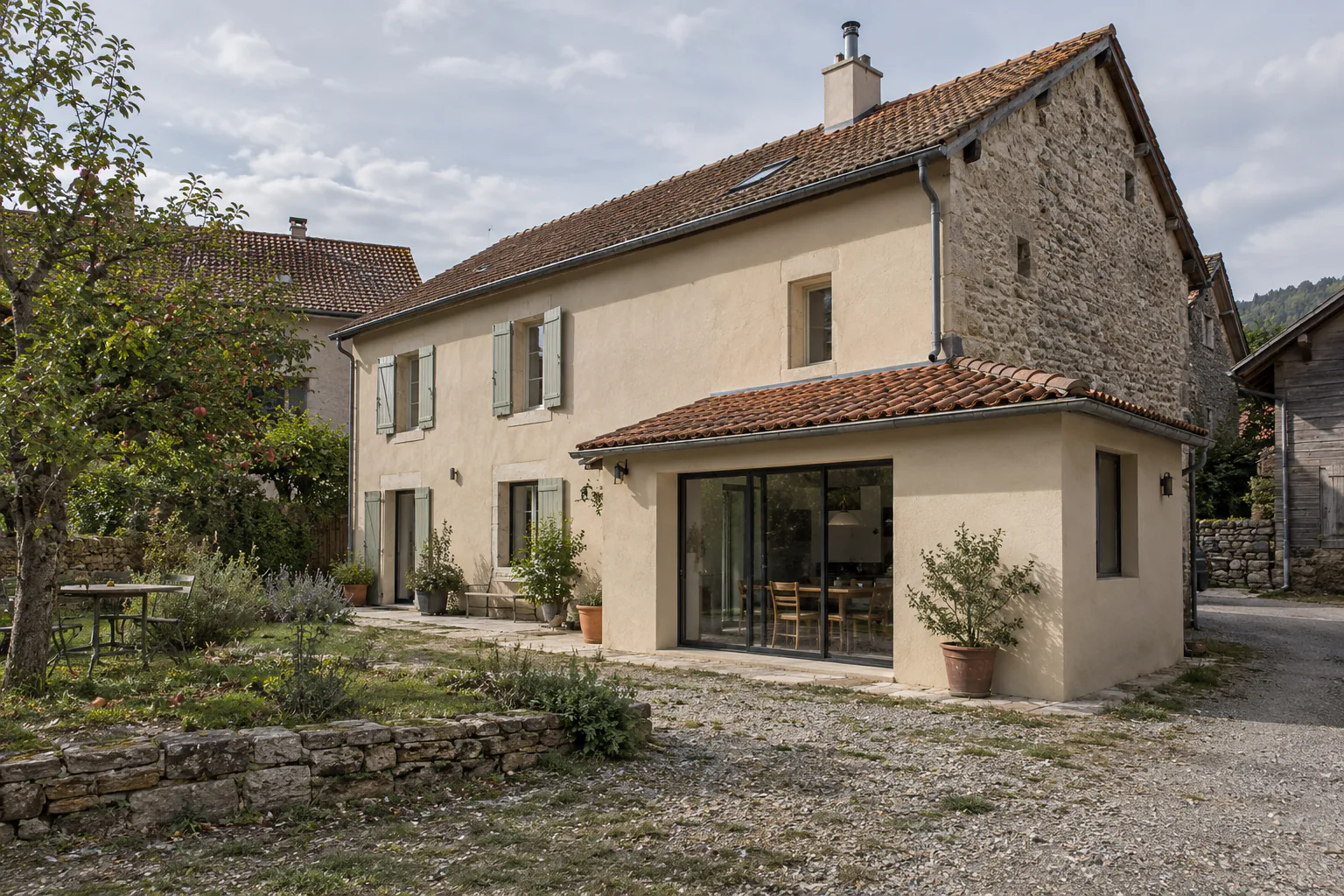 Maison ancienne et extension basse sous tuiles Une petite extension enduite à baie vitrée et toiture en tuiles rejoint une maison ancienne devant une cour de gravier.
