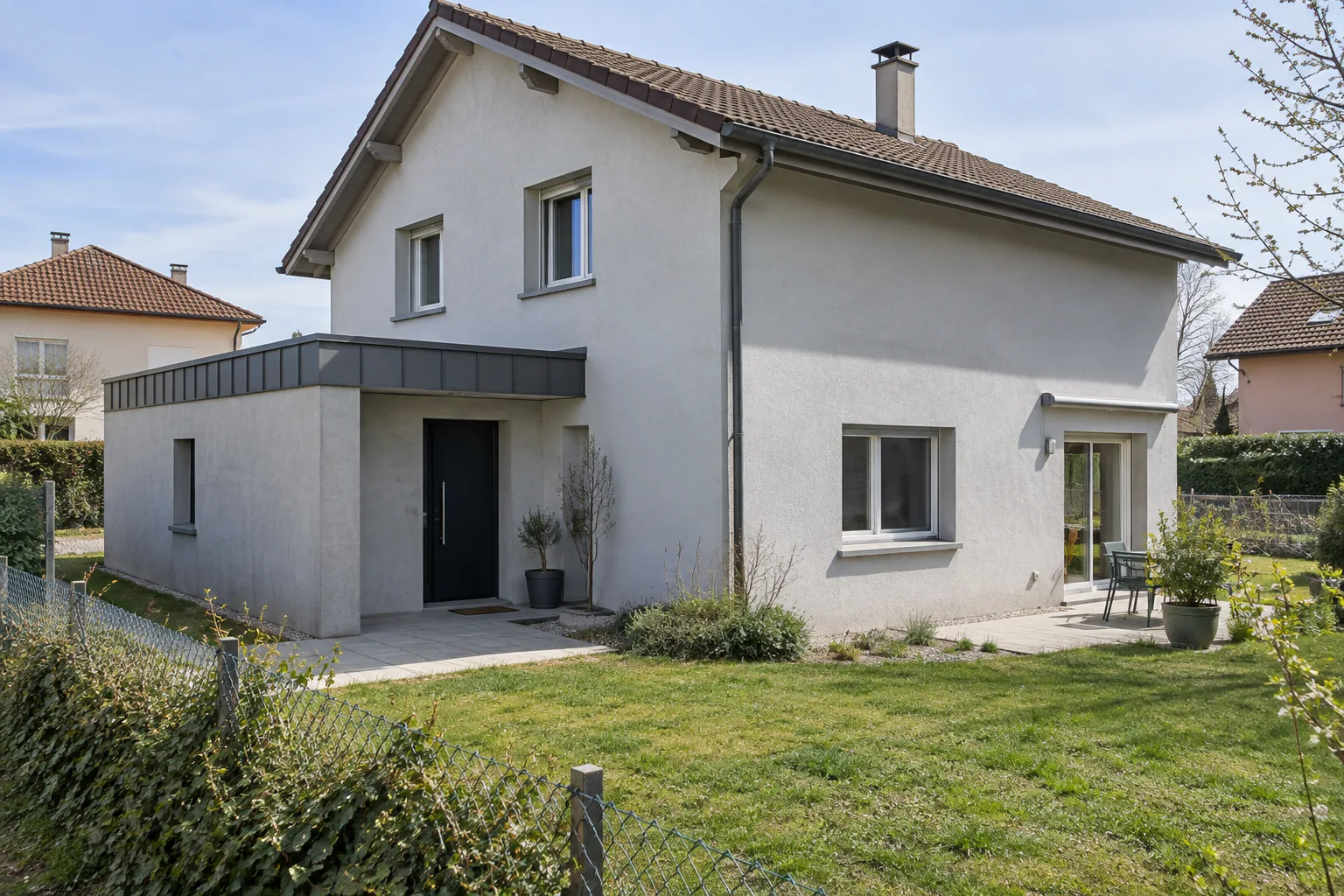 Maison blanche et entrée basse à toit métallique Un petit volume clair à toiture métallique abrite l’entrée d’une maison blanche, au bord d’une pelouse.