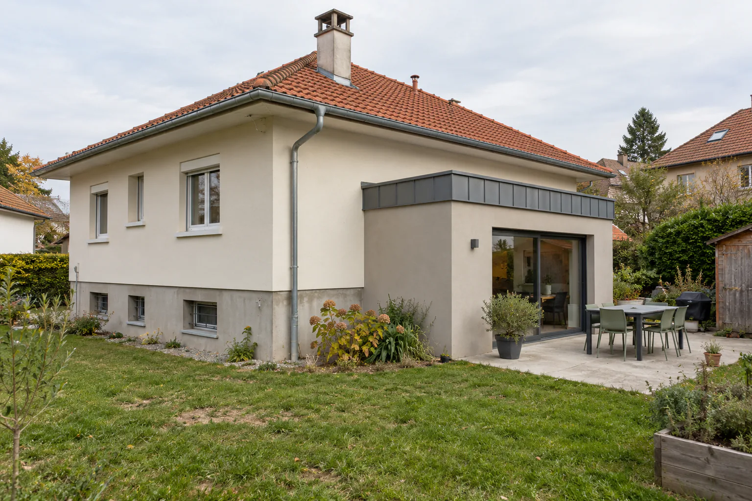 Pavillon et extension claire à couronnement métallique Une extension claire à grande baie vitrée rejoint un pavillon, devant une terrasse avec table et une pelouse.