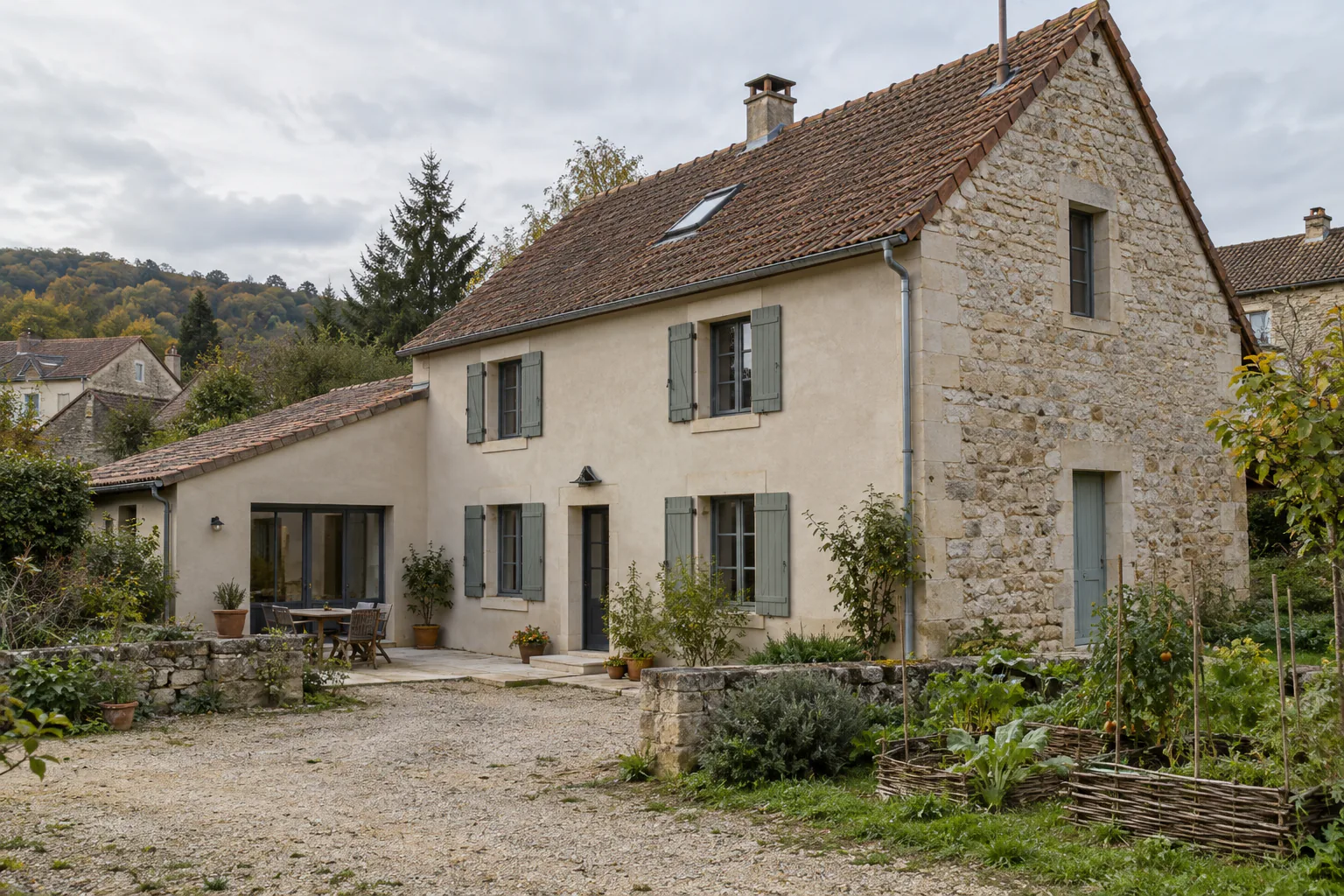 Maison en pierre et annexe claire sur cour Une maison ancienne en pierre présente une annexe basse claire, des volets gris et une cour de gravier plantée.