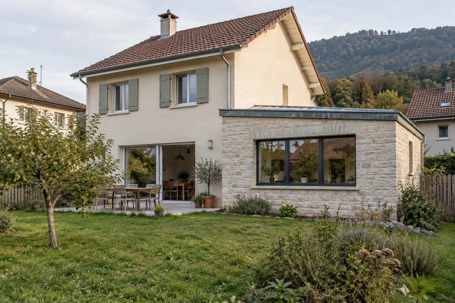Maison claire et extension en pierre sur jardin Une extension basse à parement de pierre et large fenêtre prolonge une maison claire aux volets gris.