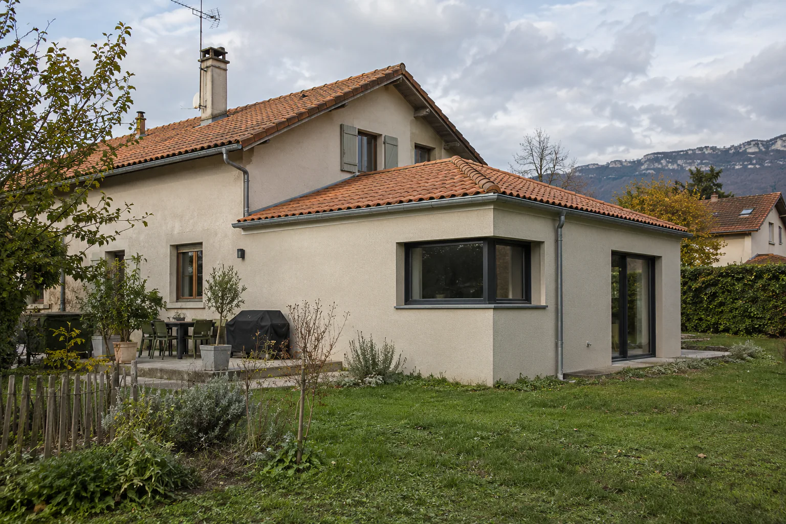 Extension claire avec fenêtre d’angle sous tuiles Un volume de plain-pied sous tuiles possède une fenêtre d’angle et une porte vitrée, devant le jardin d’une maison.