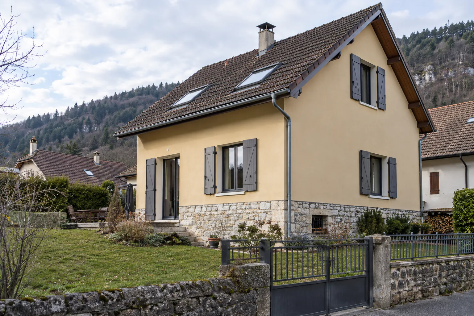 Maison ocre pâle et volets gris derrière un portail Une maison à toiture brune et fenêtres de toit présente des volets gris, au-dessus d’un soubassement de pierre et d’un jardin.