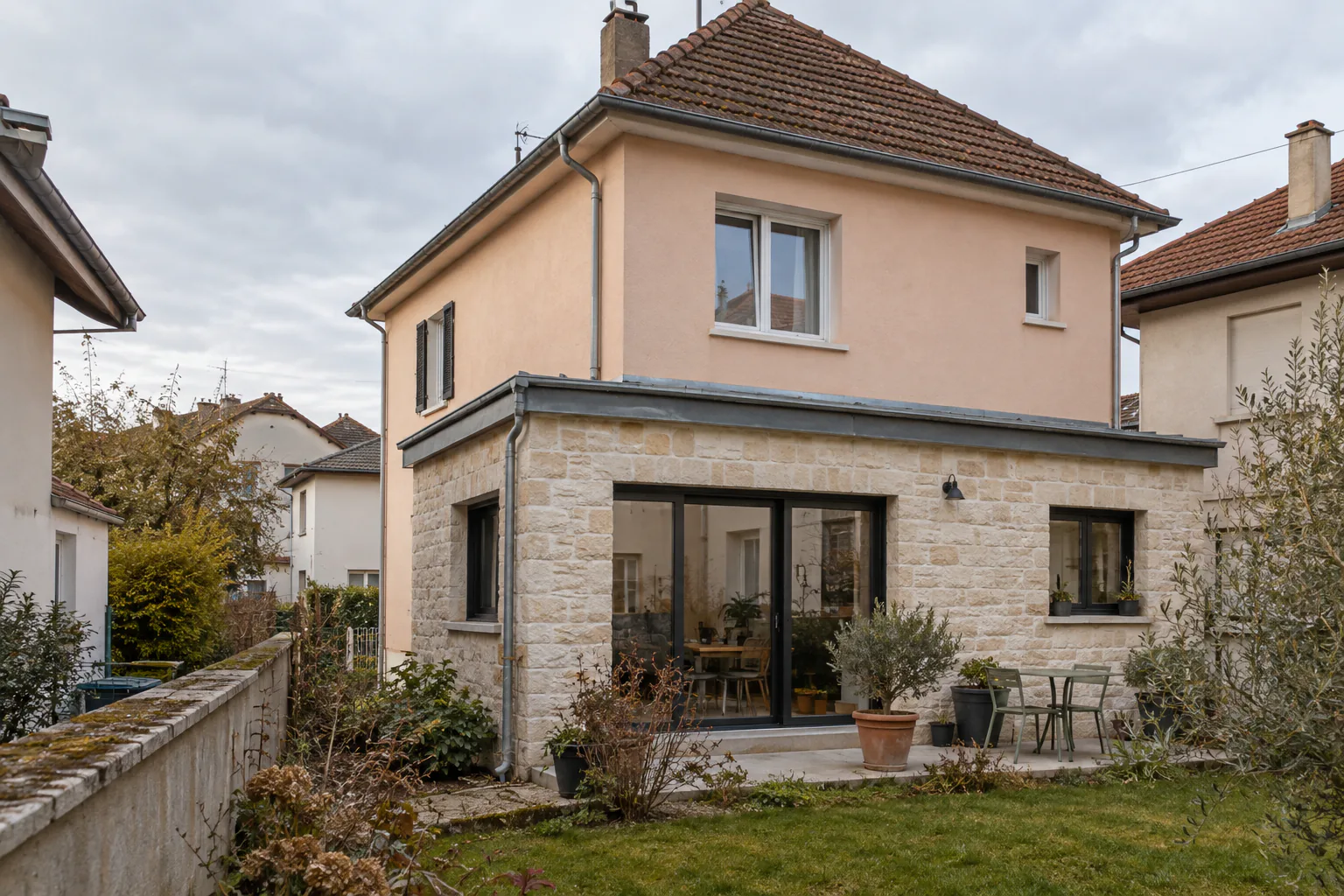 Maison rose et extension basse à parement de pierre Une extension en pierre à baie vitrée et petite fenêtre prolonge une maison rose à toiture à quatre pans.