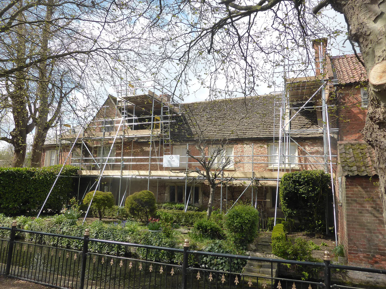 Maison et toiture derrière des échafaudages Maison entourée d'échafaudages pour des travaux de toiture, derrière des arbres et une clôture.