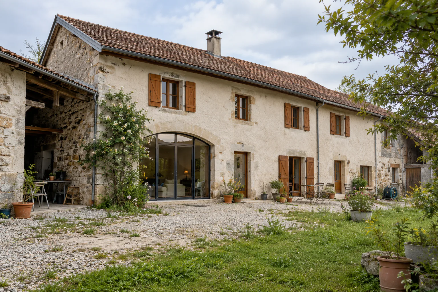 Longue maison en pierre et cour gravillonnée Long bâtiment en pierre et enduit clair avec volets en bois et grande baie cintrée.