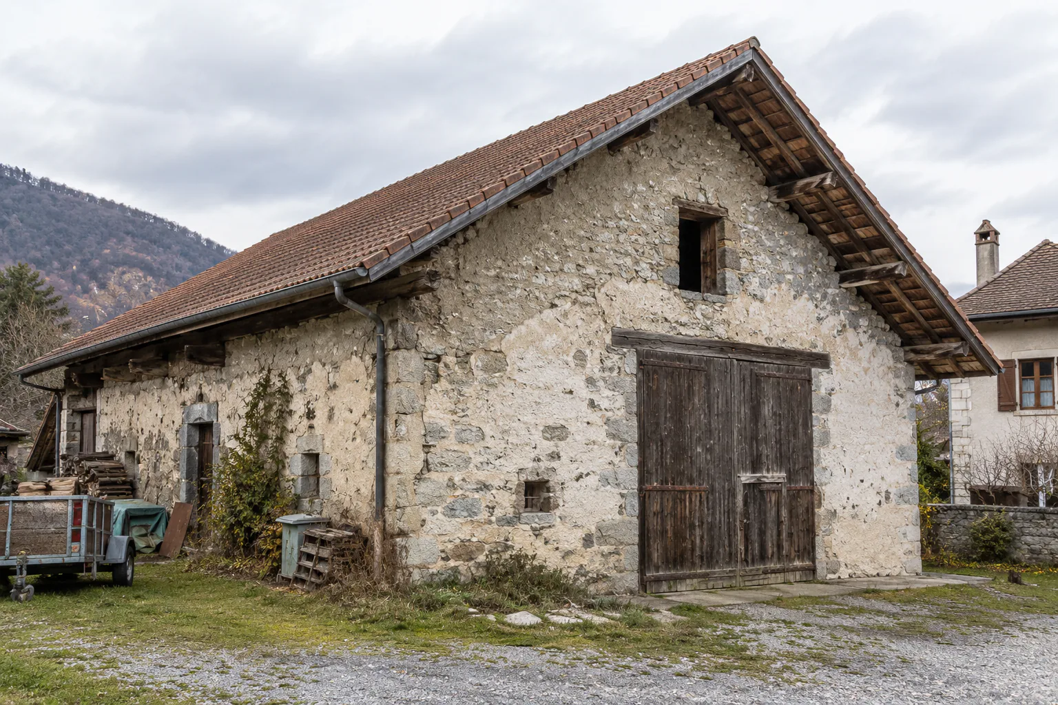 Pignon de grange en pierre brute Grange en pierre avec grande porte en bois, petite ouverture haute et toit en tuiles.
