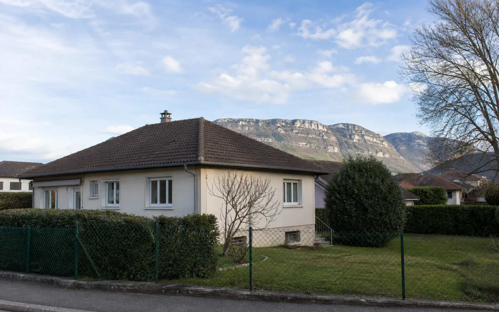 Maison basse avec montagne en arrière-plan Maison claire de plain-pied avec toiture brune, haies et relief montagneux derrière.