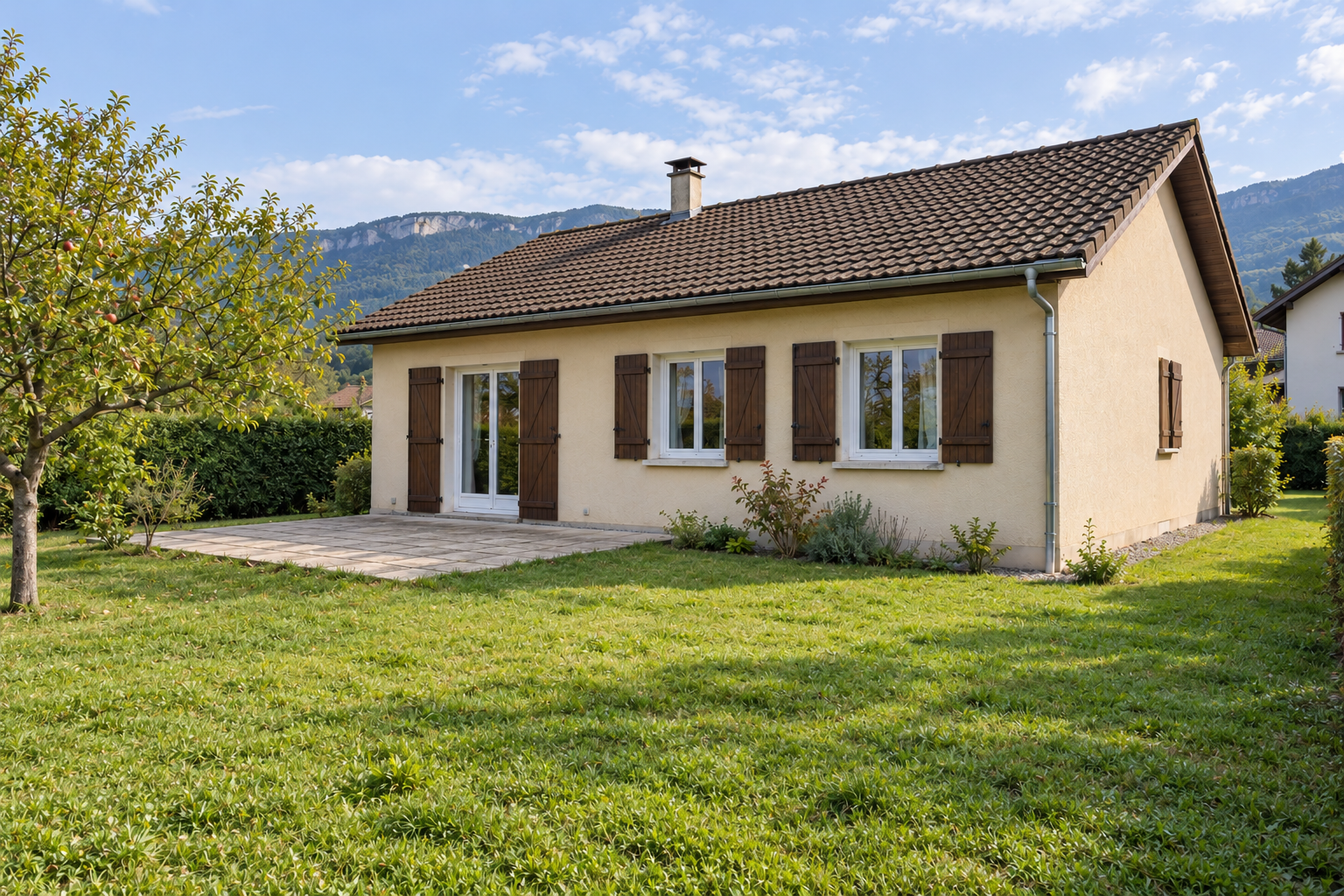 Maison de plain-pied enduite crème, toit en tuiles brunes, porte-fenêtre à gauche, deux fenêtres à volets bruns à droite et jardin arboré.